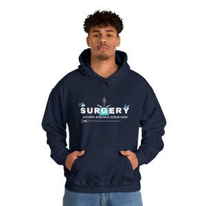 Può includere: Felpa con cappuccio blu navy con la scritta "SURGERY" in bianco e azzurro, insieme a illustrazioni di strumenti medici. Il testo "SUTURES, SCALPELS, SCRUB CAPS" è in basso. La felpa ha una tasca frontale e un cappuccio.