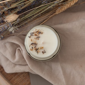 Peut inclure: Une bougie ronde blanche avec un bord noir et blanc, ornée de lavande séchée et d'autres éléments botaniques. La bougie est posée sur un tissu beige, avec de la lavande séchée et des épis de blé en arrière-plan.
