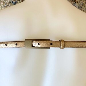 May include: A thin, metallic gold belt featuring a subtle snakeskin-like embossed texture throughout the strap. The belt includes a matching gold-toned rectangular buckle, a single fixed loop, and a series of evenly spaced adjustment holes.