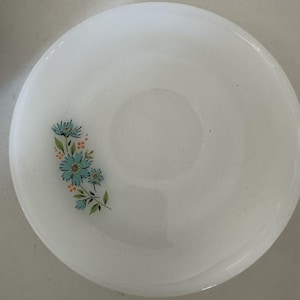 以下が含まれることがあります： 白い丸いお皿。水色の花とオレンジ色のアクセント、緑の葉のフローラルデザイン。お皿はわずかに縁が盛り上がっており、表面は滑らかです。