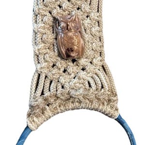 May include: A macrame towel holder with a blue ring at the top and bottom. The beige macrame has a woven pattern and features a brown owl-shaped embellishment in the center.
