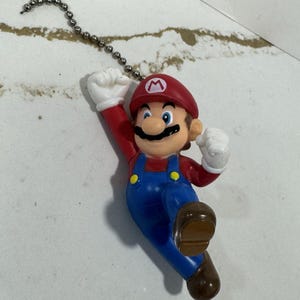 Puede incluir: Una figura de plástico de Mario, personaje de la serie de videojuegos Super Mario. Mario lleva un gorro rojo con una "M" blanca, una camisa roja de manga larga, un peto azul y zapatos marrones. La figura está unida a una cadena plateada.