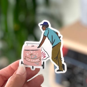 May include: A sticker of a person pushing a pink ice cream cart with the words "King's Ice Cream" on the side. The person is wearing a blue shirt and a blue hat.