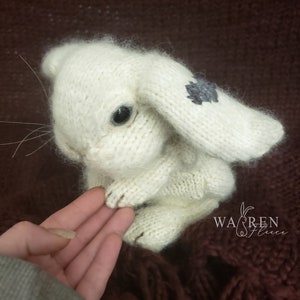May include: White knitted bunny rabbit with a gray embroidered flower on its ear. The bunny is sitting on a hand.