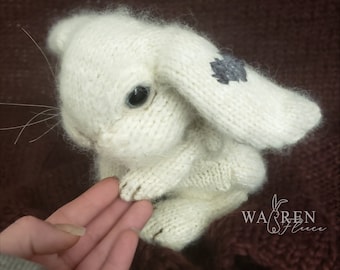 May include: White knitted bunny rabbit with a gray embroidered flower on its ear. The bunny is sitting on a hand.