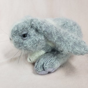 May include: A gray knitted bunny rabbit plush toy with white accents. The bunny is lying on its side with its head turned to the left.