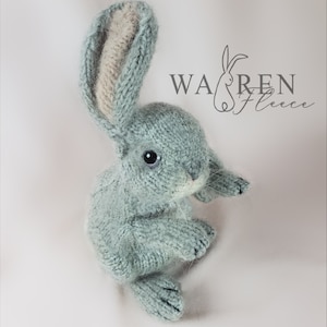 May include: A hand-knitted plush bunny toy in a light blue color with a beige inner ear. The bunny is sitting up with its ears perked up. The toy is made by Warren Fleece.