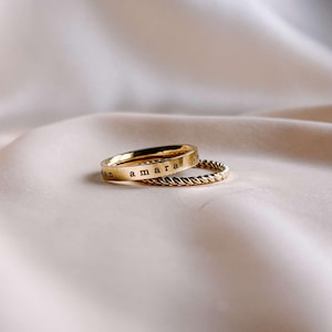 May include: Two gold rings stacked on top of each other. The top ring is a thin band with the word "amara" engraved on it. The bottom ring is a thicker band with a twisted rope design.
