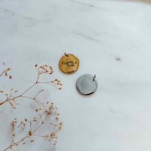 May include: Two round pendants, one gold-toned and one silver-toned, rest on a white marble surface. The gold pendant features the inscription "Rosemary Beach '26" and the name "Jeanie." Both pendants have small loops for attaching to a chain.