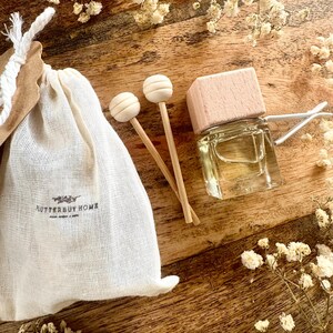 May include: A clear glass bottle with a light pink wooden cap, a small white drawstring bag with the text "Flutterby Home", and two wooden diffuser sticks with round tops. The items are arranged on a wooden surface with dried flowers.