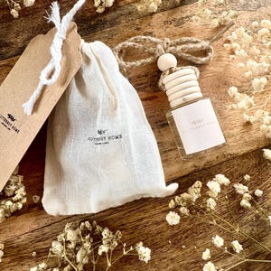 May include: Aromatherapy set comprising a small glass bottle with a wooden cap, a drawstring bag, and a tag. The bottle is labelled "Flutterby Home." The set is on a wooden surface, with dried white flowers.