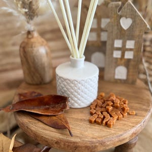 May include: A white ceramic diffuser bottle with a textured design, filled with reeds and sitting on a wooden stand. The diffuser is surrounded by brown leaves and caramel sweets.