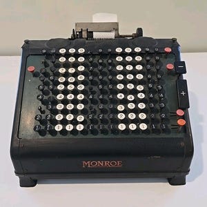 Op de afbeelding: Vintage Monroe mechanische rekenmachine met zwarte toetsen en een donkergroene en zwarte behuizing. De rekenmachine heeft meerdere rijen numerieke toetsen, een papieren rol en een plus- en min-toets. Het woord "MONROE" staat op de voorkant.