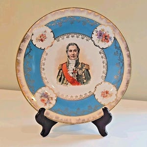 May include: A decorative porcelain plate featuring a portrait of a man in military attire, set against a light blue and white background. The plate has a gold rim and floral accents. The plate is displayed on a black stand.