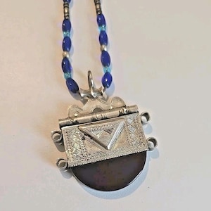 May include: A silver pendant necklace with a dark, semi-circular stone. The pendant has intricate geometric designs and a triangular accent. The necklace features a beaded strand of blue and silver beads.