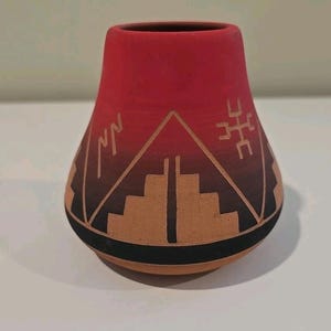 May include: A handcrafted, red and brown ceramic vase with a unique design. The vase features a geometric pattern with a black base and a gradient from red to brown. The design includes angular shapes and lines, creating a Southwestern aesthetic.