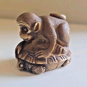 May include: A detailed sculpture of a monkey atop a turtle. The monkey is brown with a detailed face and is curled up on the turtle's back. The turtle is also brown with a textured shell. The sculpture is on a white surface.