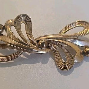 May include: A vintage gold-toned brooch with an abstract design. The brooch features swirling, ribbon-like shapes and small, spherical accents. The metal has a slightly aged appearance, suggesting it is an antique or vintage piece. The brooch is a decorative accessory.