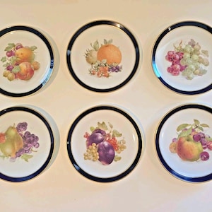 Puede incluir: Un juego de seis platos redondos de porcelana blanca, cada uno con una ilustración de frutas coloridas en el centro. Los platos tienen un borde ancho de color azul marino oscuro, acentuado por una fina línea dorada en los bordes.