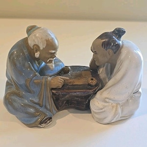 May include: A ceramic figurine of two elderly men engaged in a board game. One figure, in a blue robe, points at the board, while the other, in a white robe, observes. The board is brown with small, round game pieces.