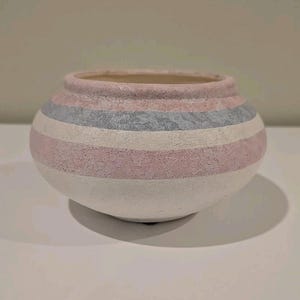 Puede incluir: Un jarrón redondo de cerámica con superficie texturizada. El jarrón presenta rayas horizontales en blanco, rosa claro y gris. El interior del jarrón es de color beige claro. El jarrón está sobre una superficie blanca.