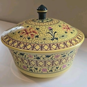 May include: A yellow ceramic lidded serving bowl with floral and geometric patterns. The bowl features pink, red, and green floral designs, with a dark green finial on the lid. The rim has a repeating geometric pattern in red and brown.