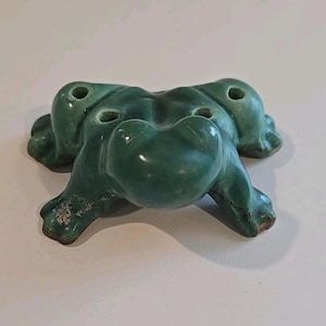 May include: A ceramic frog figurine in a glossy, jade green glaze. The frog has a rounded body, four stubby legs, and three holes on its back. The figurine is a decorative item.
