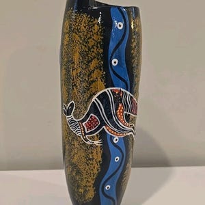 May include: A black vase with an Aboriginal art design. The vase features a kangaroo illustration in white, red, and orange, set against a blue river-like pattern and a textured gold and black background.