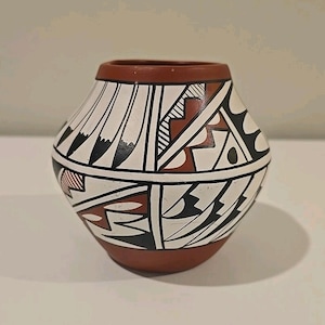 May include: A hand-painted ceramic pot with a rounded body and a wide opening. The pot features a white background with geometric designs in black, red, and brown. The rim and base are brown.