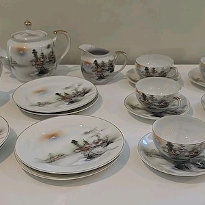 May include: A vintage porcelain tea set featuring a landscape design. The set includes a teapot, creamer, sugar bowl, plates, and teacups with saucers. The design depicts a mountain scene in grey, brown, and gold. The set is displayed on a white surface.