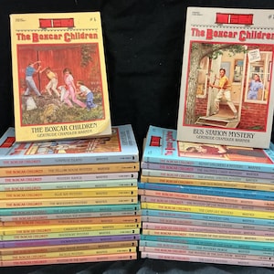 May include: A collection of The Boxcar Children paperback books by Gertrude Chandler Warner, stacked in two columns. The top books feature illustrated covers with titles including The Boxcar Children and Bus Station Mystery, with various pastel-colored spines.