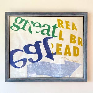 May include: A framed print with the words "great bread" in green, yellow, and blue. The print includes the number "69" in blue and a blue banner with text. The frame is a muted blue.
