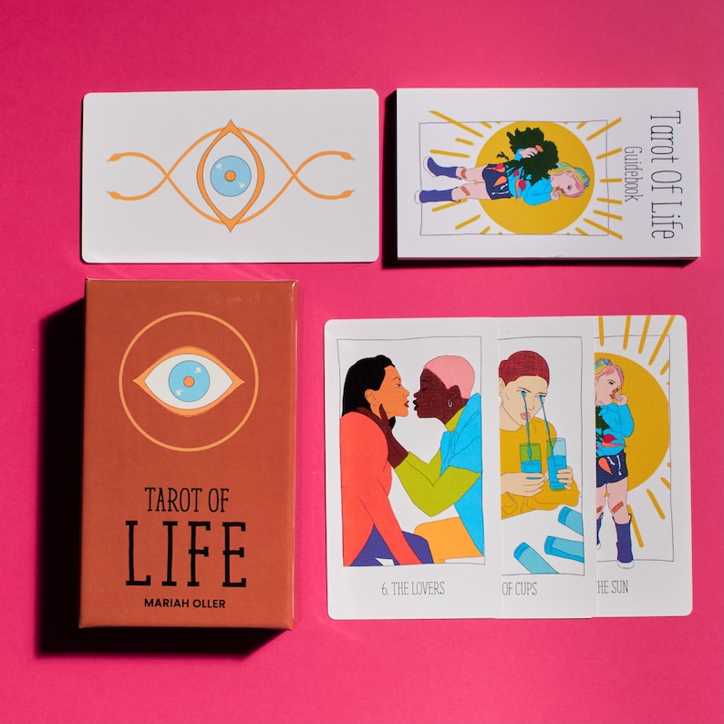 K&ouml;nnte beinhalten: Tarot of Life Kartendeckel mit braunem Hintergrund und einem gro&szlig;en Augensymbol. Auf der Schachtel steht der Text "Tarot of Life" und "Mariah Oller". Die Kartenschachtel befindet sich neben einem Satz von drei Tarotkarten mit Illustrationen eines Paares, einer Person, die eine Tasse h&auml;lt, und einer Person, die in der Sonne steht.
