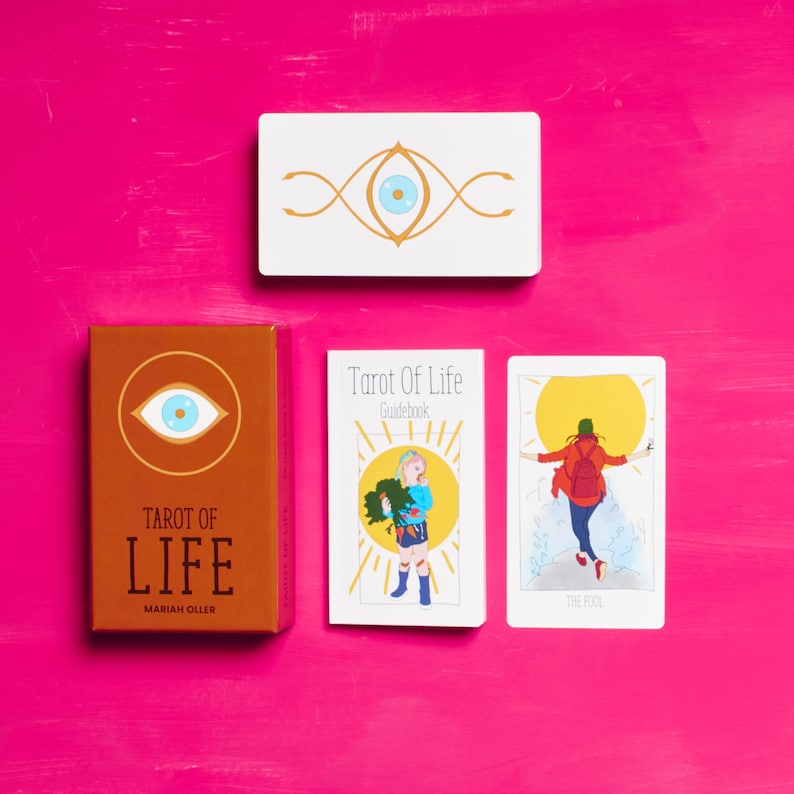 K&ouml;nnte beinhalten: Tarot of Life Deck mit einer braunen Schachtel, die ein blaues Auge mit goldenen Akzenten zeigt. Das Handbuch ist wei&szlig; mit einer gelben Sonne und einem Kind, das eine gr&uuml;ne Pflanze h&auml;lt. Eine Karte aus dem Deck zeigt eine Person in rot-blauer Kleidung mit einem Rucksack, die auf einem Felsen steht, mit einer gelben Sonne hinter ihr. Der Kartentext lautet "The Fool".