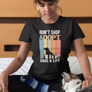 Pu&ograve; includere: T-shirt nero con la scritta "DON'T SHOP, ADOPT, SAVE A LIFE" in un design retr&ograve;. Il design include una silhouette di cane e impronte di zampe. La maglietta &egrave; indossata da una persona seduta su un letto con un gatto.