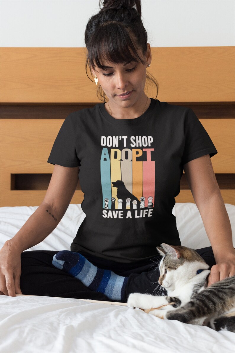 Pu&ograve; includere: T-shirt nero con la scritta "DON'T SHOP, ADOPT, SAVE A LIFE" in un design retr&ograve;. Il design include una silhouette di cane e impronte di zampe. La maglietta &egrave; indossata da una persona seduta su un letto con un gatto.