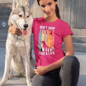 Pu&ograve; includere: Una maglietta rosa acceso con la scritta "DON'T SHOP ADOPT SAVE A LIFE" e una grafica di una sagoma di cane e impronte di zampe. Una persona indossa la maglietta ed &egrave; seduta accanto a un cane husky grigio e bianco su una superficie di cemento.