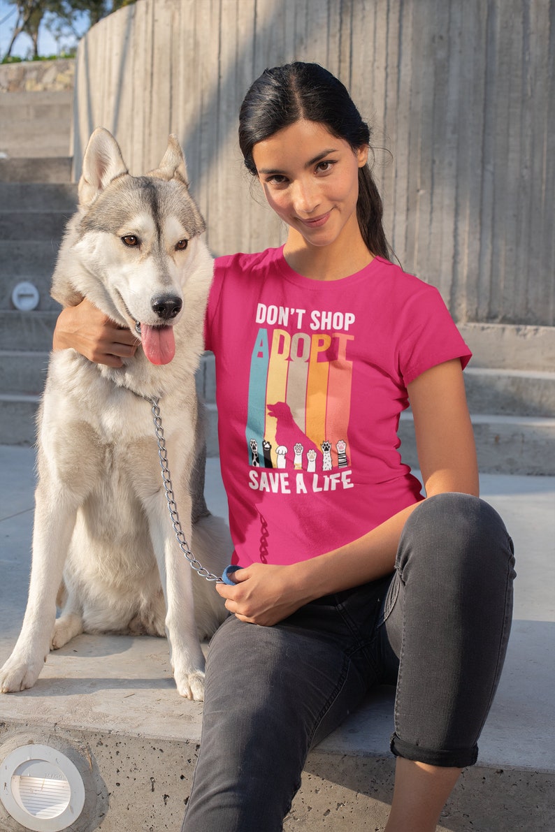 Pu&ograve; includere: Una maglietta rosa acceso con la scritta "DON'T SHOP ADOPT SAVE A LIFE" e una grafica di una sagoma di cane e impronte di zampe. Una persona indossa la maglietta ed &egrave; seduta accanto a un cane husky grigio e bianco su una superficie di cemento.
