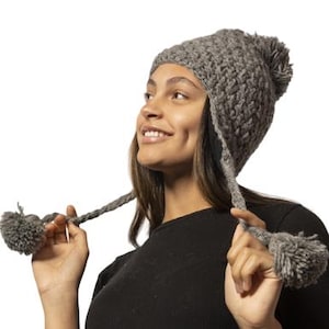 May include: A gray knitted beanie with ear flaps and pom poms. The beanie has a crocheted texture and is worn by a person.
