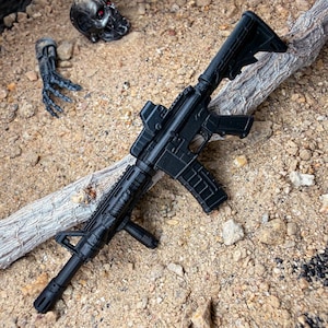 May include: A black toy rifle with a scope, positioned on a sandy surface alongside a piece of weathered wood. A skull and a robotic arm are also visible, hinting at a science fiction or action theme.