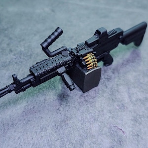 May include: A black miniature machine gun with a handle, sight, and ammunition belt. The gun is detailed with various components and a dark finish. The ammunition is gold-coloured. The object is set against a grey surface.