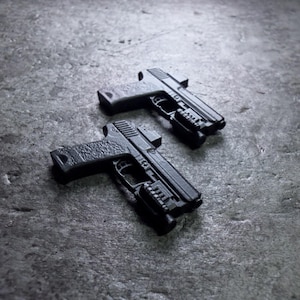 May include: Two black handguns with textured grips and attached accessories are displayed on a grey surface. The firearms are positioned at a slight angle, showcasing their design and features. The image is in grayscale.