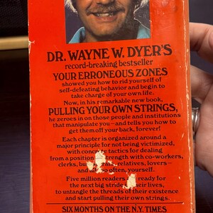 May include: Vintage paperback book cover with a portrait of Dr. Wayne W. Dyer. The cover is orange with white text, advertising "Pulling Your Own Strings" and "Your Erroneous Zones". The book is a New York Times bestseller.