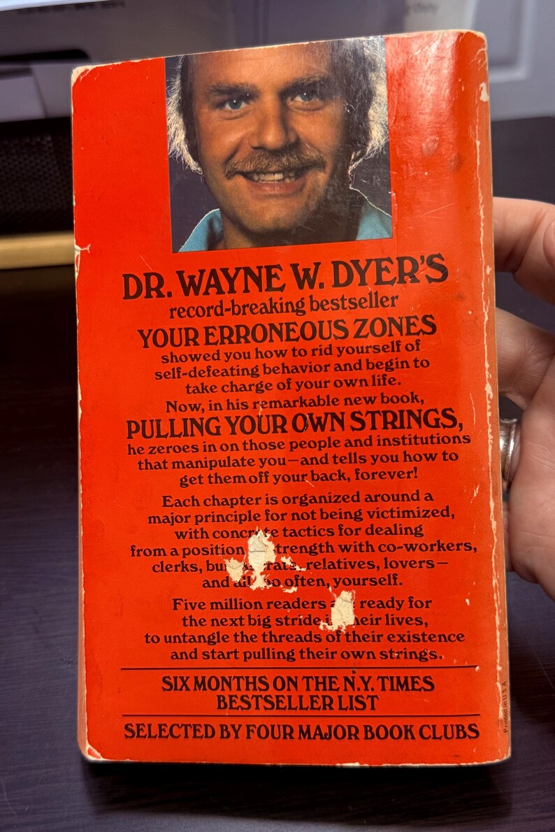 May include: Vintage paperback book cover with a portrait of Dr. Wayne W. Dyer. The cover is orange with white text, advertising "Pulling Your Own Strings" and "Your Erroneous Zones". The book is a New York Times bestseller.
