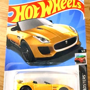 May include: A gold-colored Hot Wheels die-cast model of a '15 Jaguar F-Type Project 7. The car is displayed on a blue and orange card with the Hot Wheels logo. The package includes the text "HW Roadsters" and "3+".