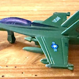 Puede incluir: Un avión de juguete verde con una capota morada y el texto "USAF" en las alas. El avión de juguete tiene insignias de estrellas azules y blancas y está hecho de plástico. El juguete es un modelo de un avión militar.