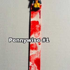 includes : A handmade bookmark features a small Pennywise figurine from the movie IT. Pennywise  #1 The IT Clown is positioned at the top of a long, rectangular bloody bookmark. The bookmark is painted white with blood splatter patterns of red blood resembling blood. The bookmark measures approximately 8 inches in length. fantastic, Stephen King, horror, fan gift or Halloween gift