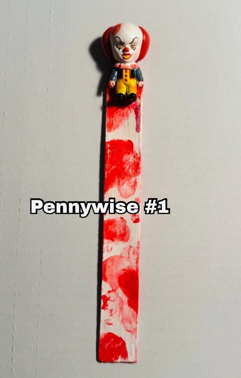 includes : A handmade bookmark features a small Pennywise figurine from the movie IT. Pennywise  #1 The IT Clown is positioned at the top of a long, rectangular bloody bookmark. The bookmark is painted white with blood splatter patterns of red blood resembling blood. The bookmark measures approximately 8 inches in length. fantastic, Stephen King, horror, fan gift or Halloween gift