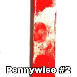 includes : A handmade bookmark features a small Pennywise figurine #2 version from the movie IT. The figurine is positioned at the top of a white bookmark with red bloody splatters resembling blood. The bookmark measures approximately 8 inches in length, as indicated. Great Stephen King horror fan gift!