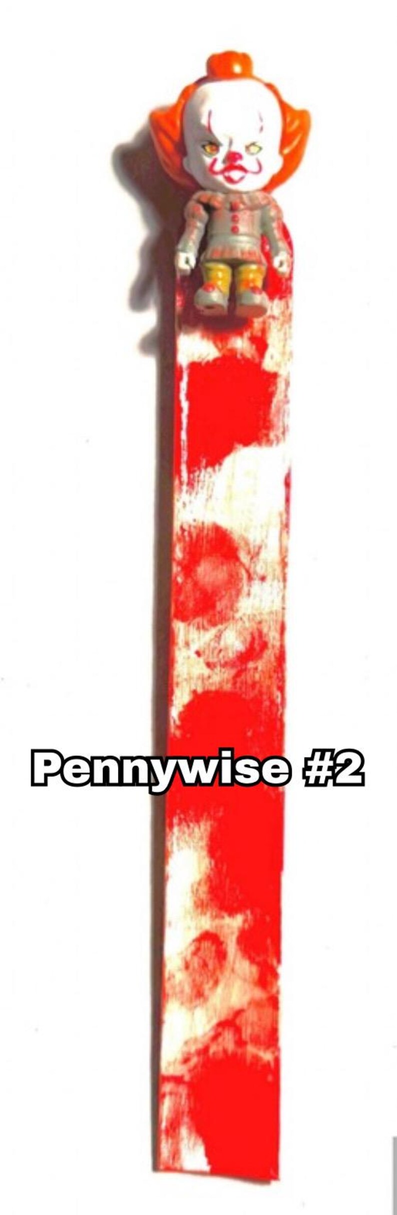 includes : A handmade bookmark features a small Pennywise figurine #2 version from the movie IT. The figurine is positioned at the top of a white bookmark with red bloody splatters resembling blood. The bookmark measures approximately 8 inches in length, as indicated. Great Stephen King horror fan gift!